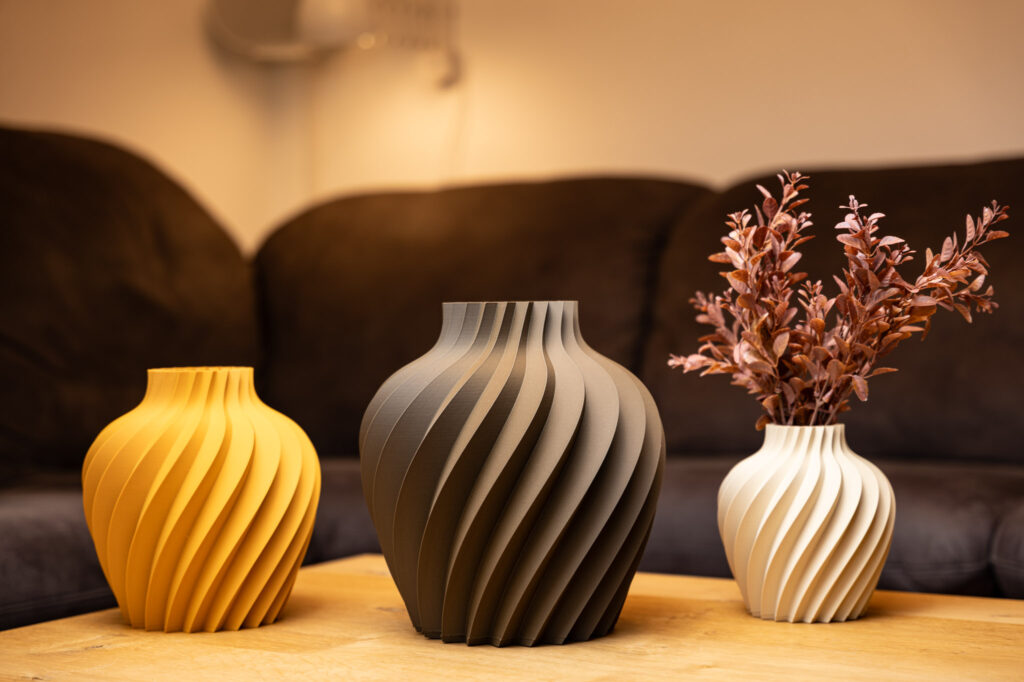 Drei stilvolle 3D-gedruckte Vasen in Gelb, Braun und Weiß mit eleganten Spiralmustern, dekorativ auf Holztisch.