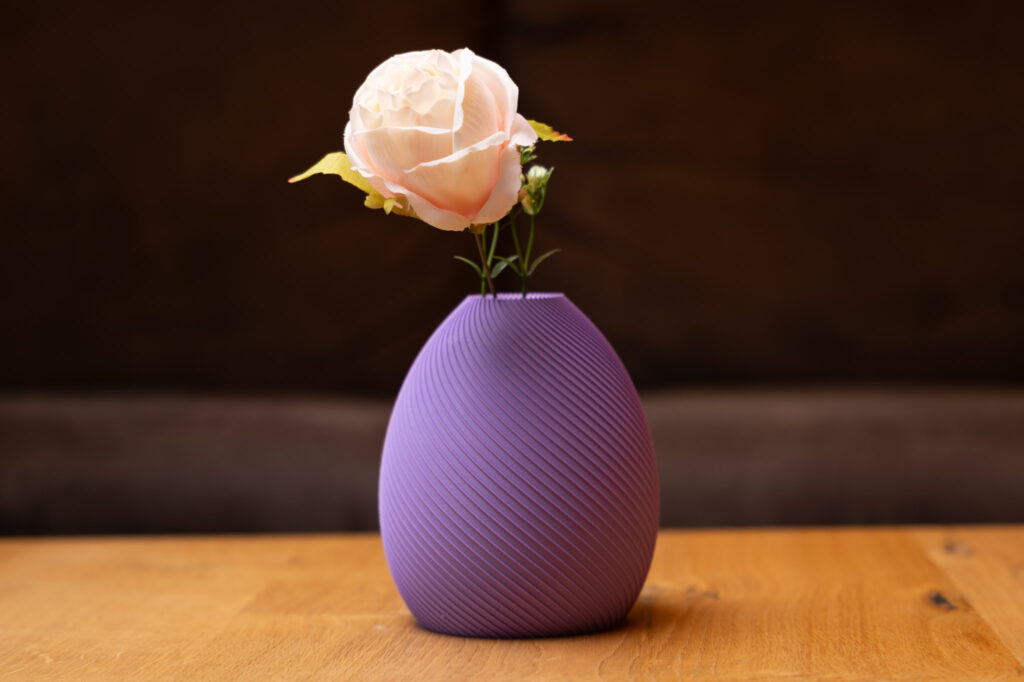 Lila 3D-gedruckte Vase in eleganter Spiralform mit zarter weißer Blume, präsentiert auf einem Holztisch, stilvoll und modern.