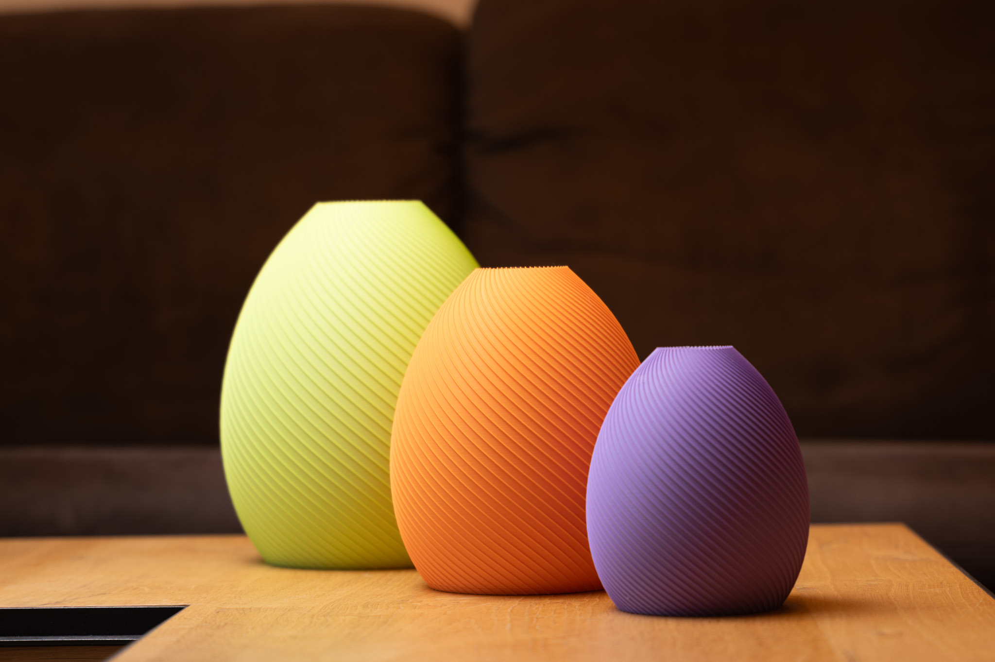 Drei elegante, farbige 3D-gedruckte Vasen in Gelb, Orange und Lila mit feiner Rillenstruktur auf Holzoberfläche.
