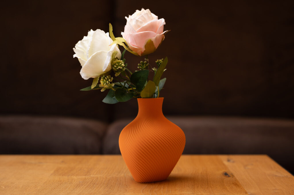 3D-gedruckte orange Vase mit eleganten Kurven, geschmückt mit einer weißen und einer rosa Blüte, auf Holztisch.