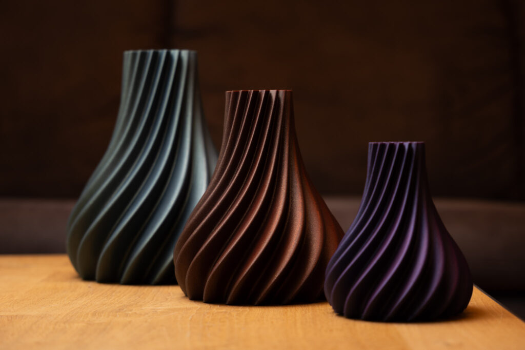 Drei stilvolle, spiralförmige Vasen aus dem 3D-Drucker in Blau, Braun und Lila auf einem Holztisch.