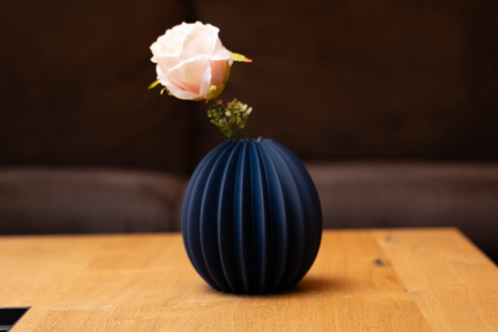 Dunkelblaue, gerippte 3D-gedruckte Vase mit eleganter rosa Blume auf hellbraunem Holztisch, stilvoll und modern.