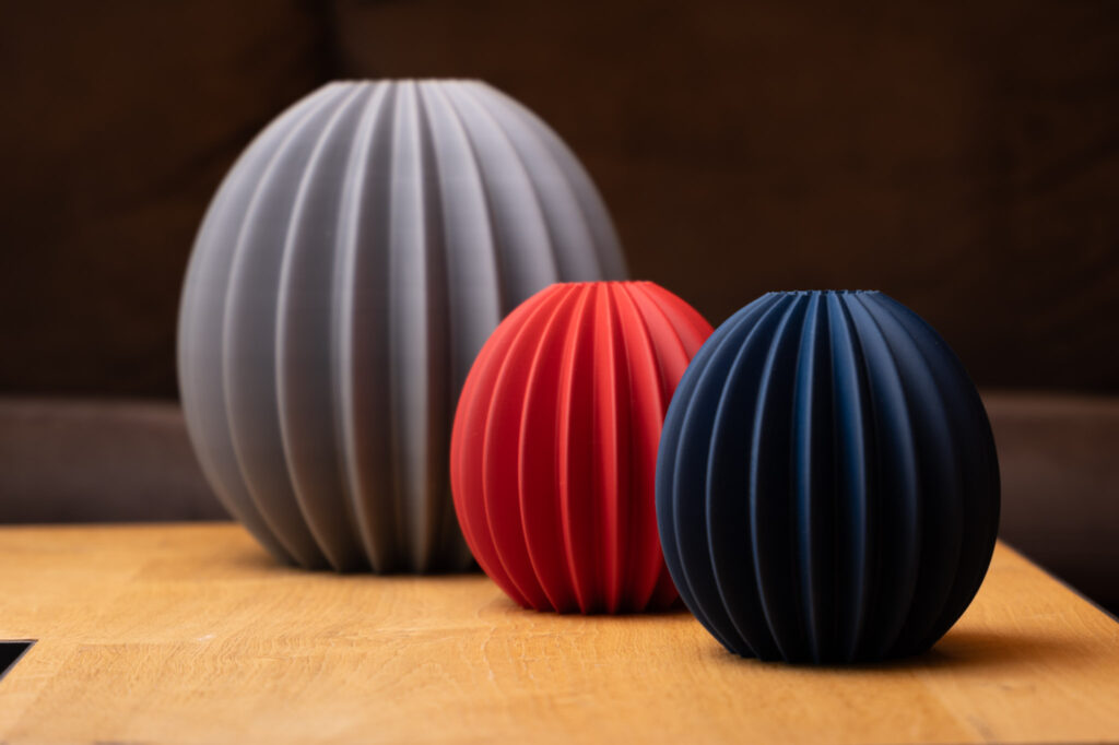 Drei moderne, geriffelte 3D-gedruckte Vasen in Grau, Rot und Blau auf einem Holztisch. Elegantes Design.