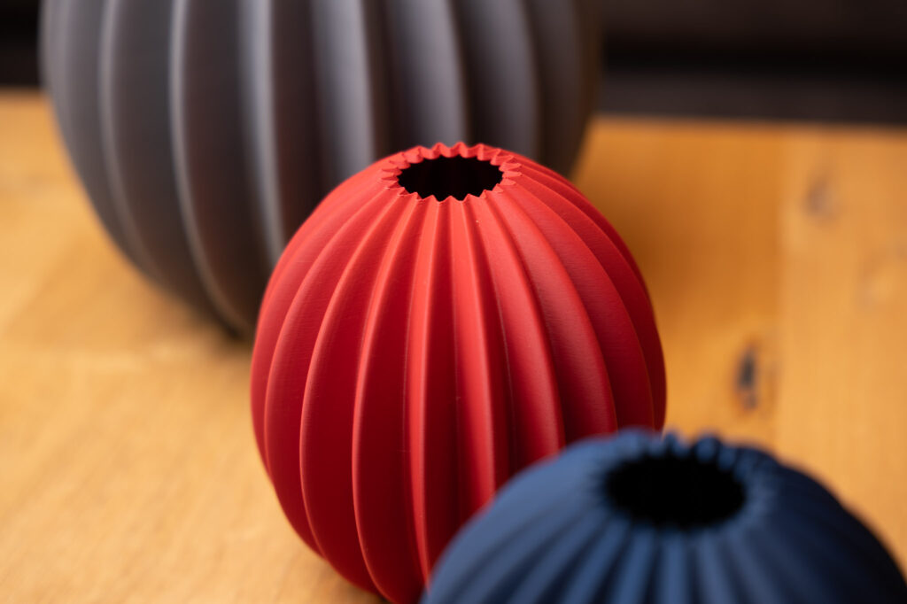 Elegante, rote 3D-gedruckte Vase mit geriffelter Oberfläche, sticht lebhaft auf hellem Holztisch hervor.