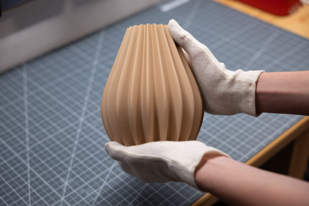 3D-gedruckte Vase mit eleganten, gerippten Formen in sanftem Beige, gehalten von behandschuhten Händen auf Schneidematte.