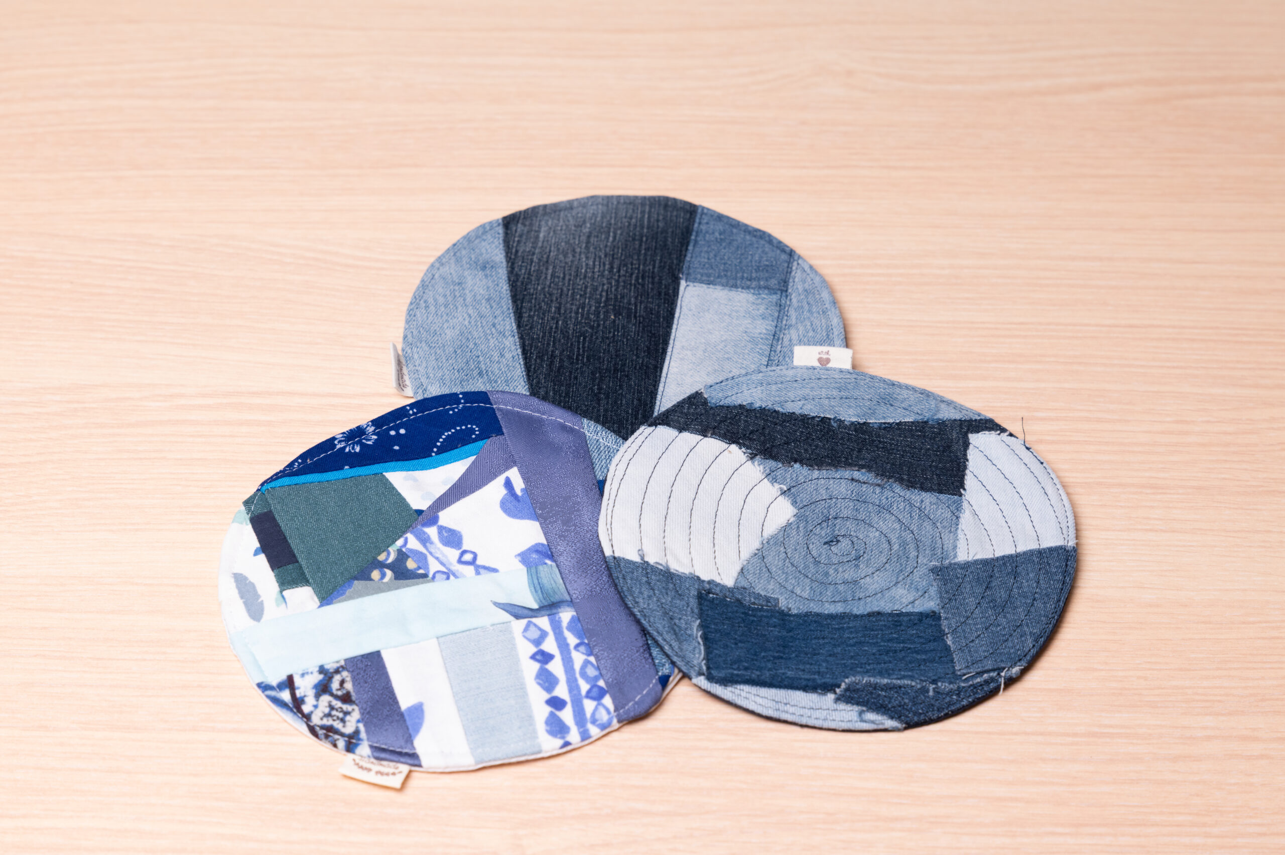 Drei stilvolle runde Patchwork-Untersetzer in Blautönen aus recyceltem Stoff auf hellem Holztisch.