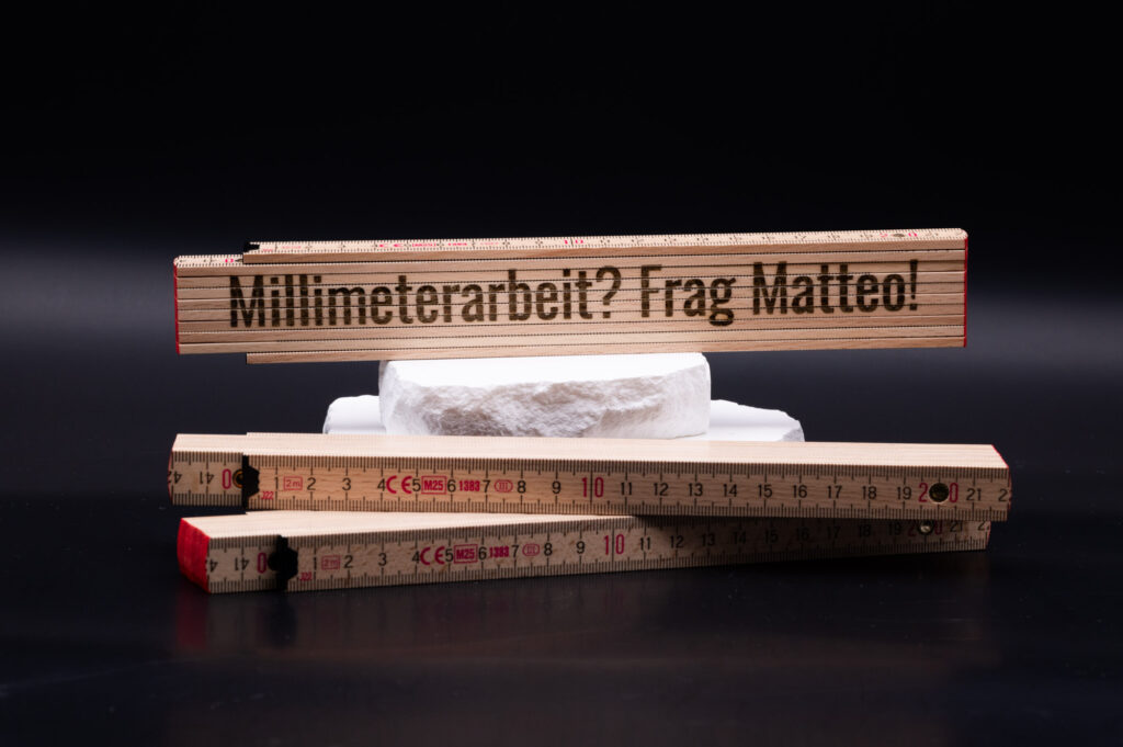 Holz-Zollstock mit Gravur Millimeterarbeit? Frag Matteo! vor einer stilvollen weißen 3D-gedruckten Plattform.