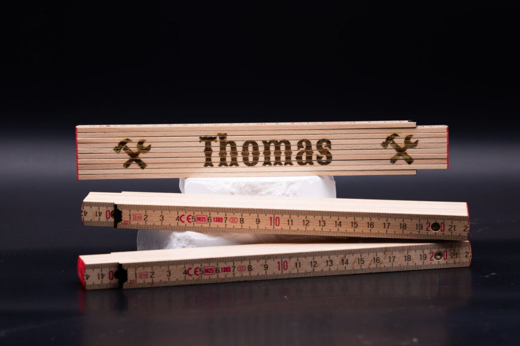 Holz-Zollstock mit eingraviertem Namen Thomas und Werkzeug-Symbolen, kunstvoll gestaltet, auf schwarzem Hintergrund präsentiert.