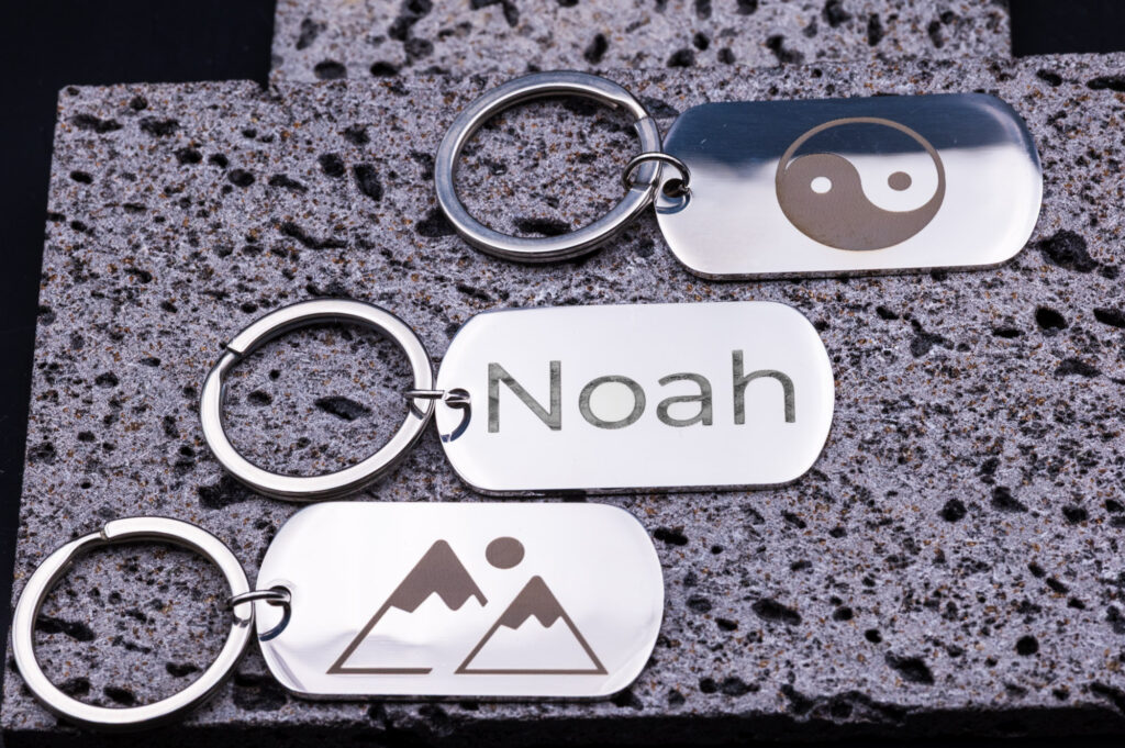 Metall-Schlüsselanhänger mit Gravuren: Name, Bergmotiv, Yin-Yang-Symbol, edel auf poröser Steinplatte präsentiert.