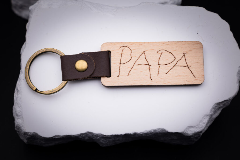 Schlüsselanhänger aus Holz mit Gravur Papa, braunem Lederriemen und goldfarbenem Ring, elegant auf weißem Hintergrund.