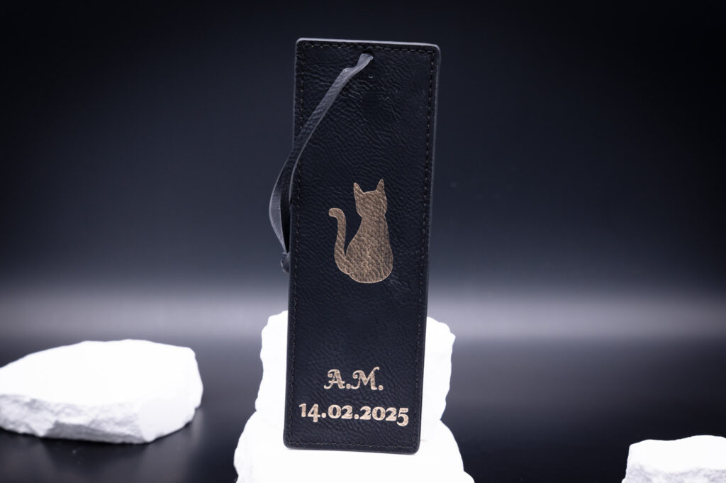 Elegantes, schwarzes Leder-Lesezeichen mit goldener Katzenprägung, personalisiert mit Initialen und Datum, vor weißem Hintergrund.