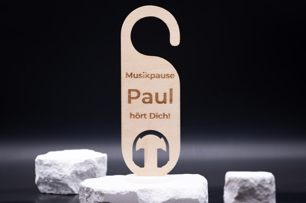 Holzschild mit Kopfhörer-Motiv und der Gravur Musikpause Paul hört Dich! vor schwarzem Hintergrund auf weißen Steinen.