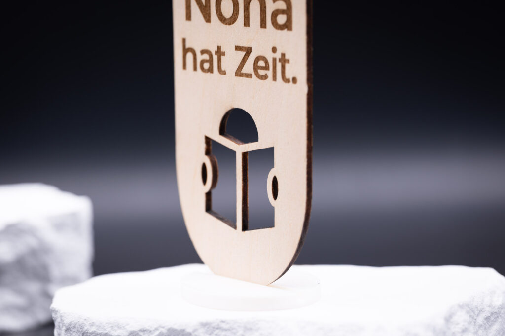 Holzschild mit elegantem Schriftzug Nona hat Zeit, kunstvoll aus hellem Holz gefertigt, vor dunklem Hintergrund.
