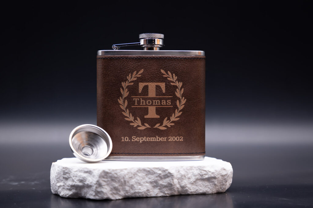 Braune Lederflasche mit graviertem Namen und Datum auf hellem Stein, edel und personalisiert präsentiert.