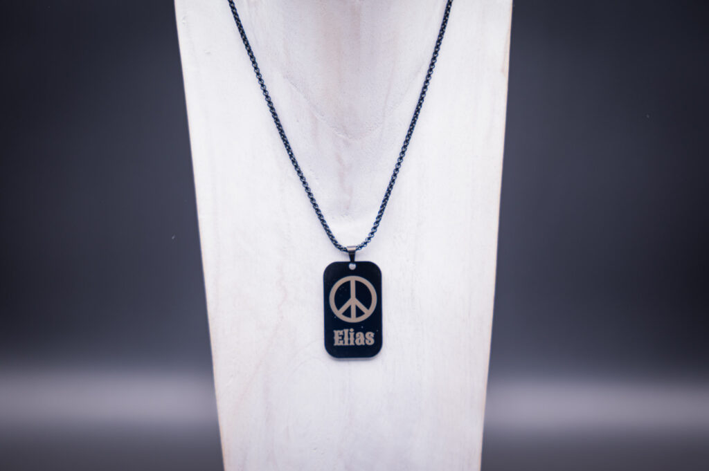 Schwarze Halskette mit Anhänger, gestaltet mit Peace-Zeichen und Name, präsentiert auf elegantem weißen Hintergrund.