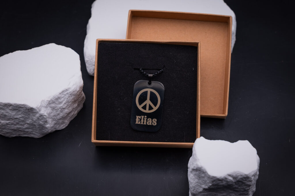 Schwarzes Schmuck-Etikett mit Peace-Zeichen und Elias-Gravur in offener Box, umgeben von weißen Steinen.