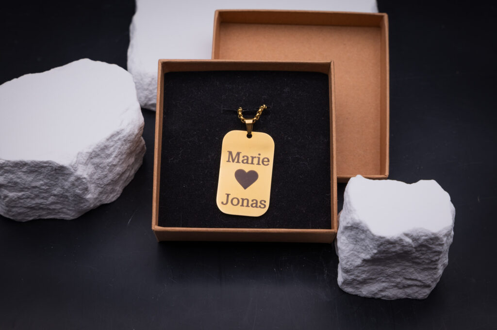 Personalisierte goldene Metallkette in Geschenkbox, graviert mit Herz und Namen, umgeben von stilvollen weißen Steinen.