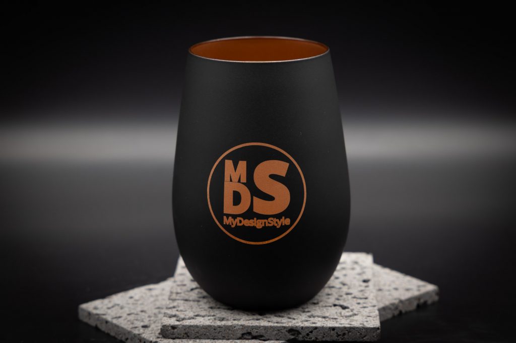 Schwarzer MyDesignStyle Becher auf grauem Stein, moderne Eleganz mit stilvollem, bronzefarbenem Logo, perfekter Designakzent.